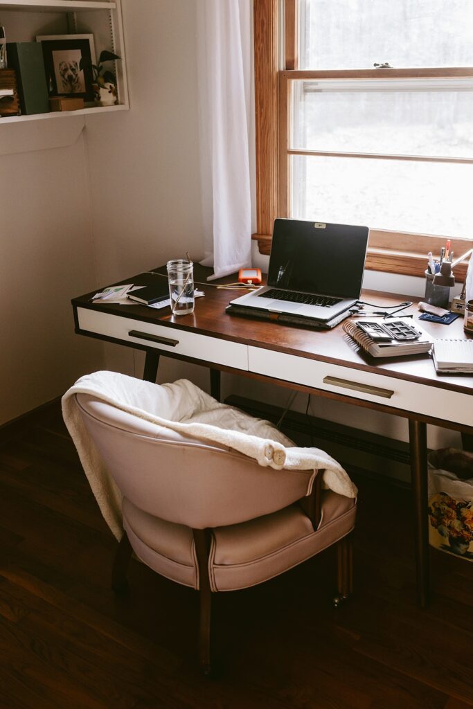 A cozy wooden desk under a window with a laptop, planner, and large glass of water on top. There's a lavender leather desk chair on brass wheels with a throw blanket on top.