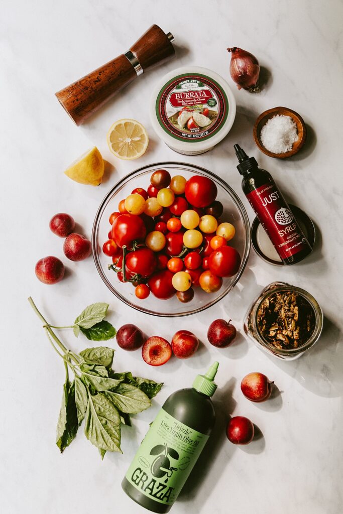 A flatlay food photograph of ingredients for Cherry Plum and Heirloom Tomato Salad. There's cherry plums, cherry tomatoes, pepita clusters, fresh basil, burrata cheese, flaky sea salt, lemon, shallot, and a pepper mill